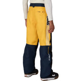 A person shown from the back wearing colorblock snow pants in yellow and navy blue, with white and navy vertical stripes on the side, paired with a white long-sleeve shirt and dark sneakers.